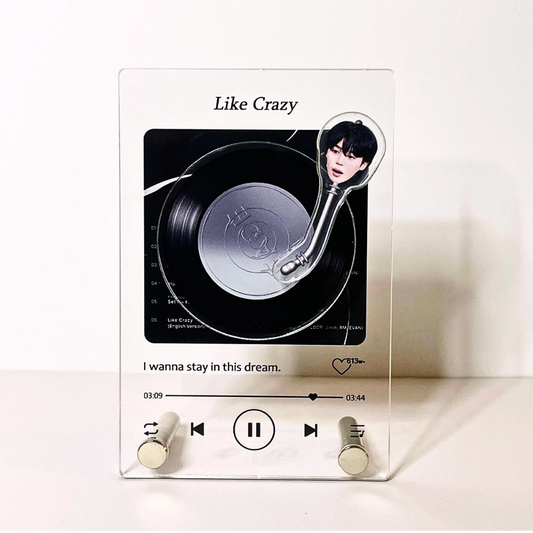 A clear acrylic music player display featuring the song 'Like Crazy' with an image of BTS Jimin. The display resembles a classic iPod, with a play/pause and forward buttons, song timeline, and additional song information in a stylized font. The text 'I wanna stay in this dream' is placed below the controls.